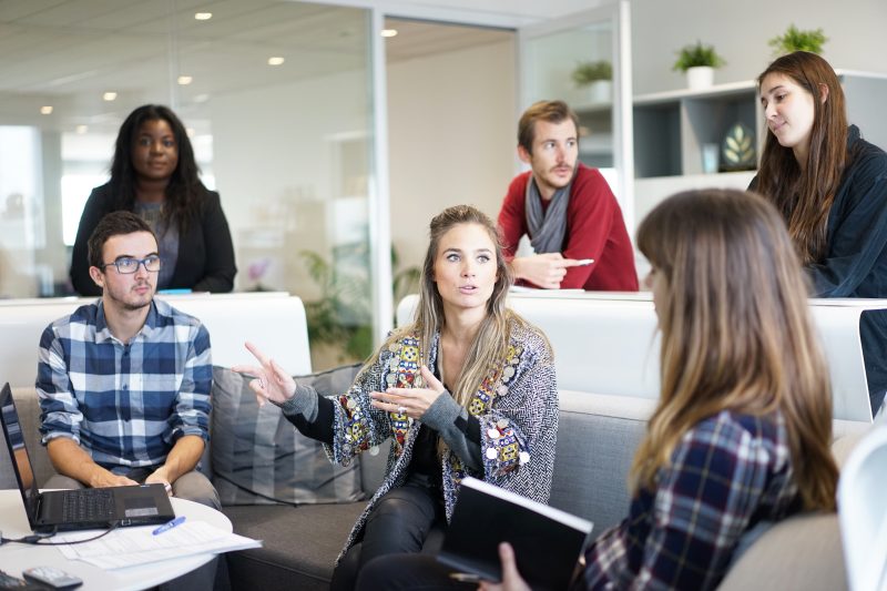 A diverse group of people, dressed business casual, sitting around a round conference room table. Six people have their hands raised, and some have smiles on their faces 