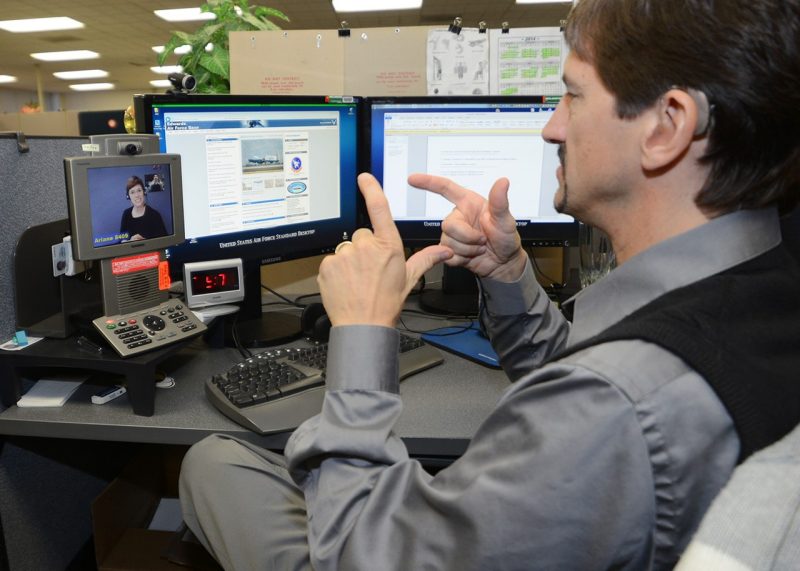 Man at desk faces video communication device, making sign language gesture